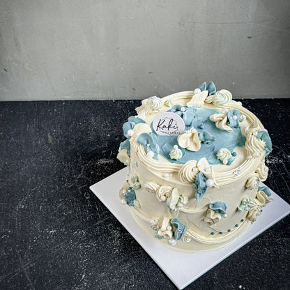 Decorative bespoke cake from Kaki Desserts with intricate frosting and colorful floral accents, displayed on a white cake stand.