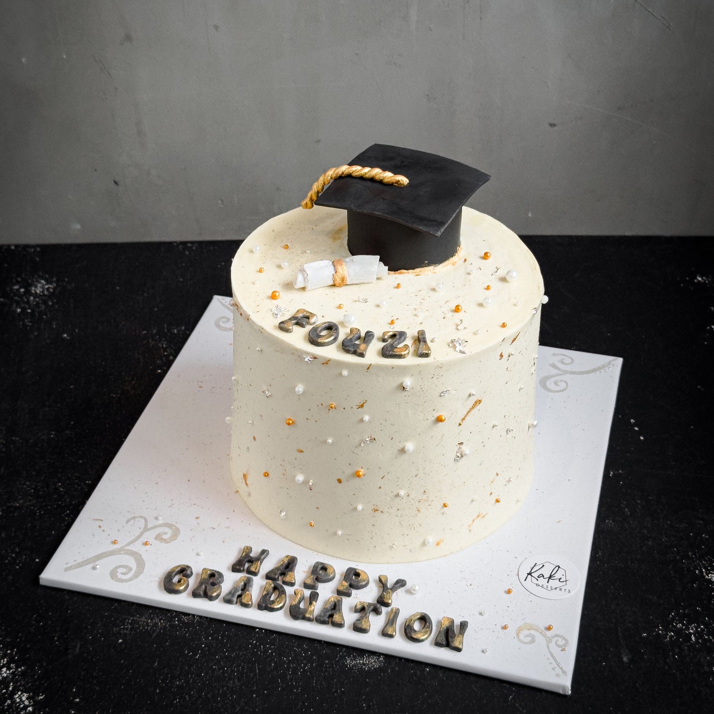 Decorative bespoke cake from Kaki Desserts with intricate icing and floral accents on a white cake stand.