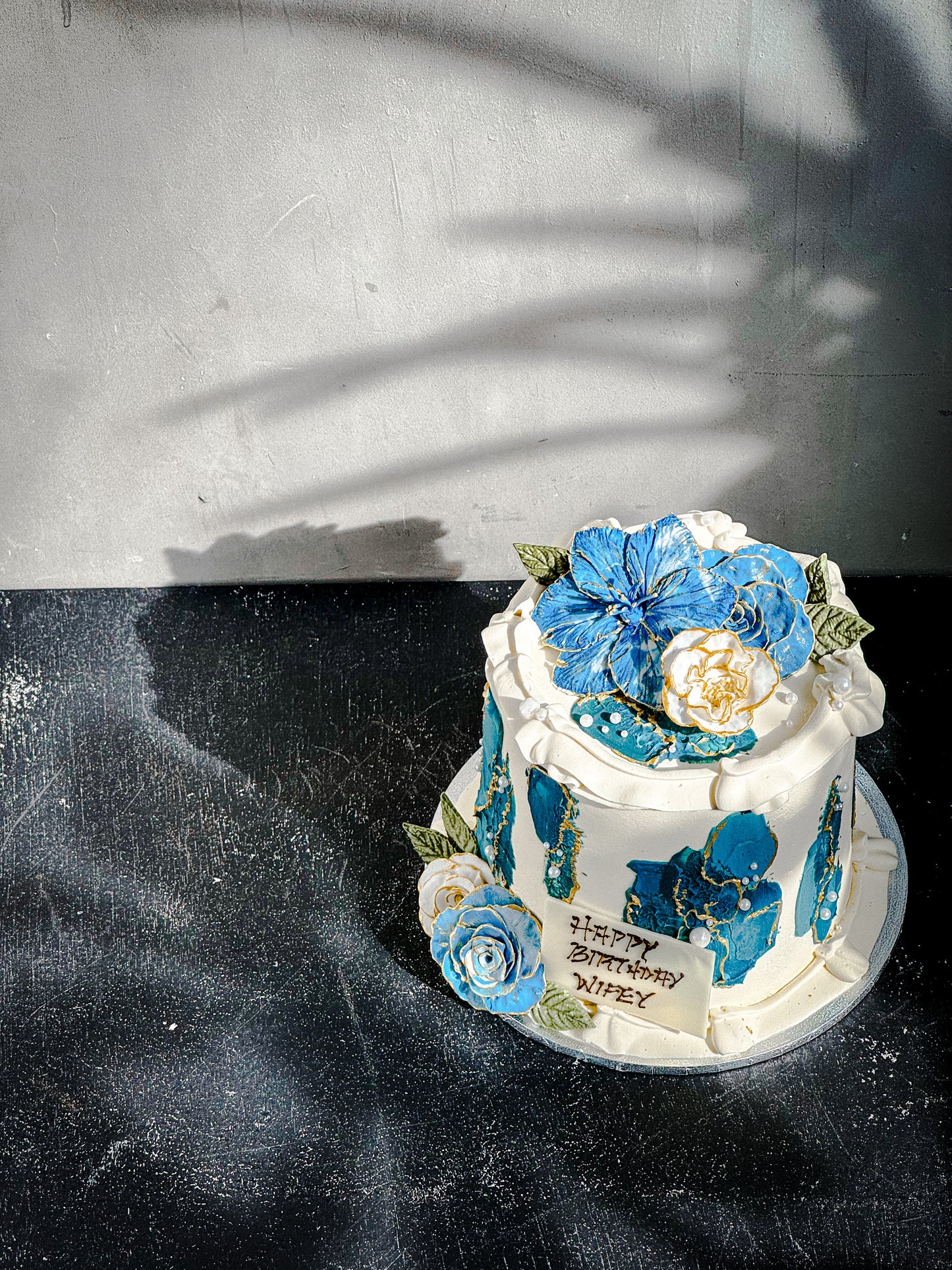 Decorative bespoke cake from Kaki Desserts with colorful floral icing and elegant piping, displayed on a white cake stand.