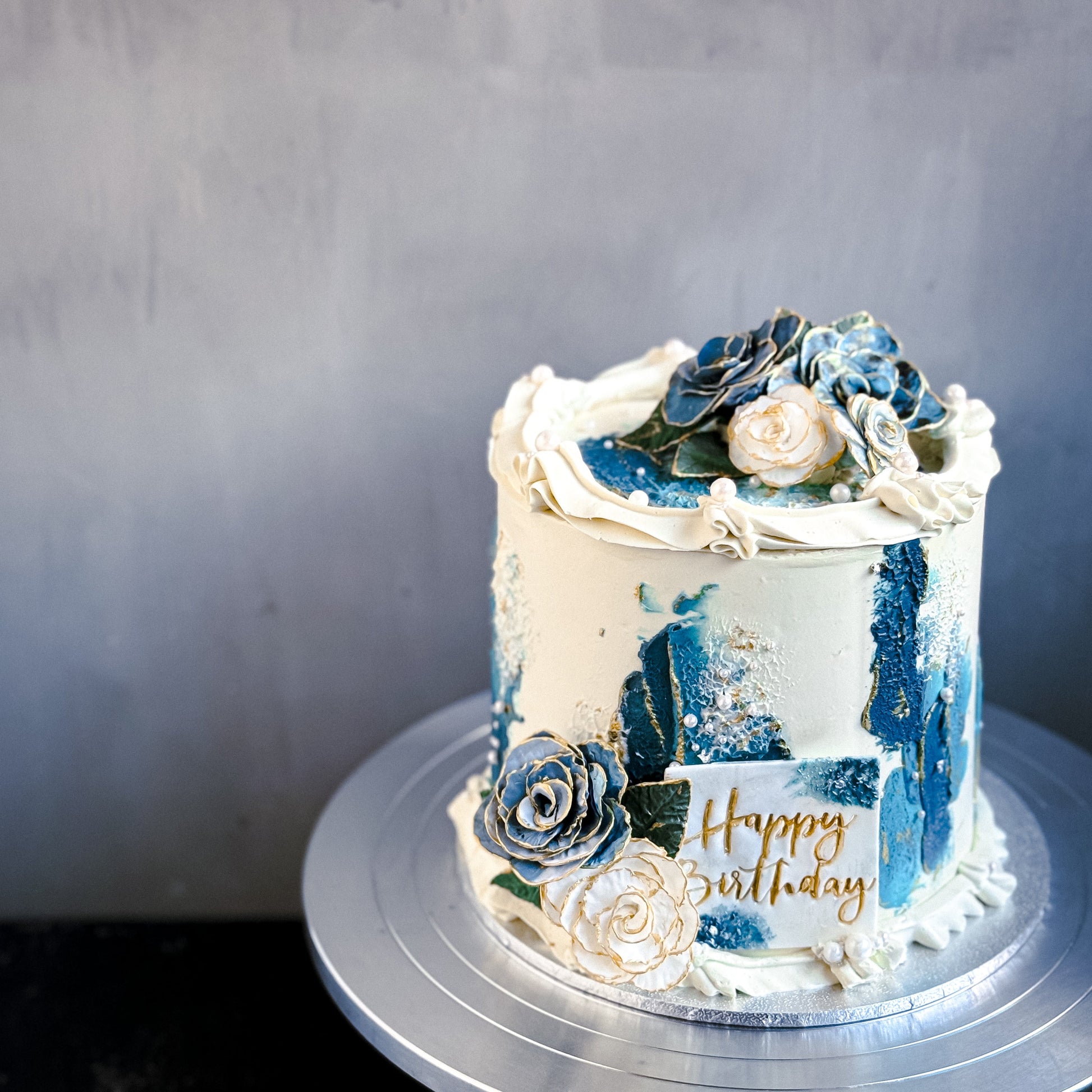 Decorative bespoke cake from Kaki Desserts with elegant floral icing and intricate piping, displayed on a white cake stand.